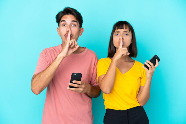 Young mixed race couple holding mobile phone isolated on blue background showing a sign of silence gesture putting finger in mouth - Photo, Image