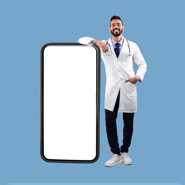 Middle Eastern Male Doctor Leaning On Big Smartphone, Blue Background, Square - Foto, Imagem