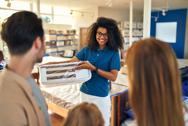 An African American woman salesperson consultant consults a young family by showing samples of mattresses in a bed, mattress and pillow store. - Φωτογραφία, εικόνα