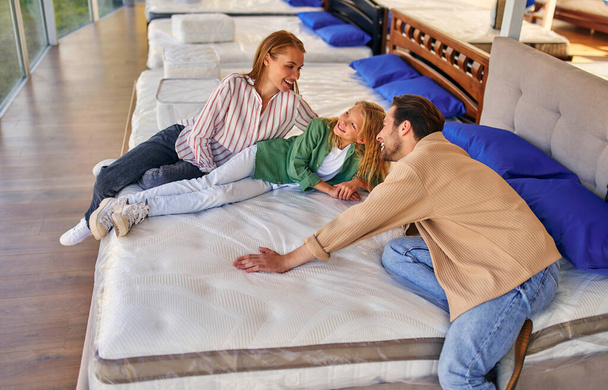 A young family with their lovely daughter lay down on the bed, trying the mattress for softness in the store. Buying a bed and bedding. - Foto, Imagen