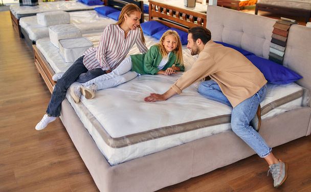 Egy fiatal család a bájos lányukkal feküdt az ágyon, próbálták a matracot puhaságért a boltban. Ágynemű és ágynemű vásárlása. - Fotó, kép