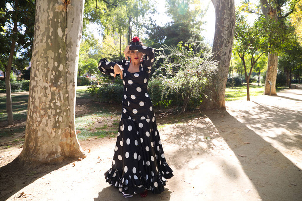 Spanyol, gyönyörű, barna flamenco táncos egy tipikus flamenco ruha fekete, fehér pöttyös. A sevillai parkban táncol. Flamenco koncepció az emberiség kulturális öröksége. - Fotó, kép