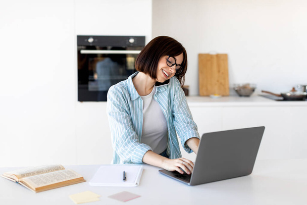 Manager job, work remotely. Happy young woman talking by cellphone and typing on laptop computer, sitting in kitchen - Foto, immagini
