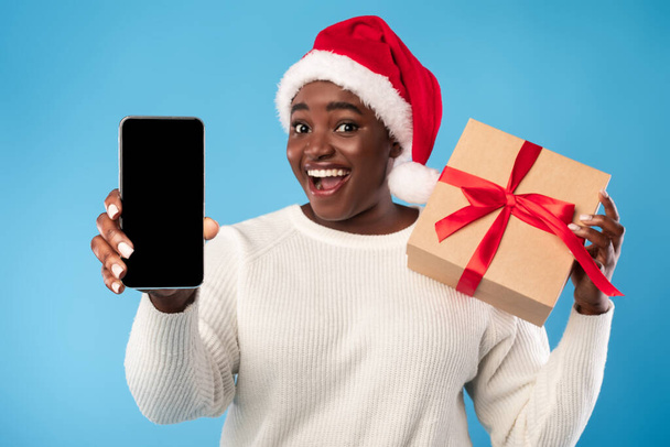 Αφρικανική γυναίκα Εμφάνιση Smartphone Screen Holding Χριστουγεννιάτικο δώρο, μπλε φόντο - Φωτογραφία, εικόνα