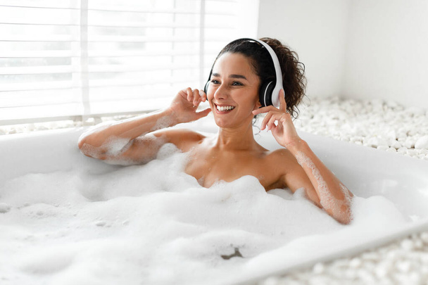 Jovem encantadora em fones de ouvido ouvindo música enquanto estava deitado em banho quente em casa, espaço livre - Foto, Imagem