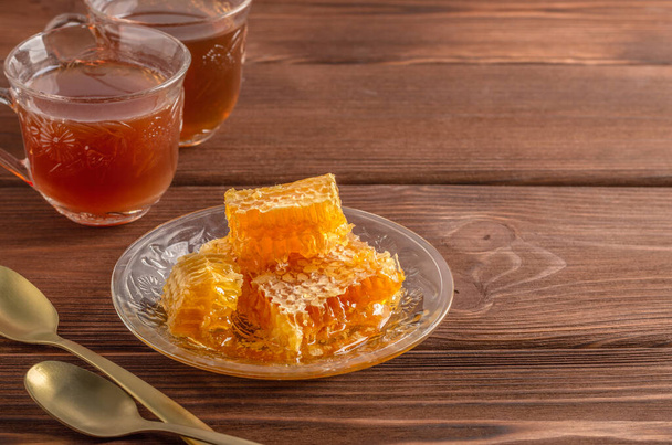 Honigwabe auf einem Glasteller und zwei Tassen Tee mit Löffeln auf einem Holztisch. Kopierraum. - Foto, Bild