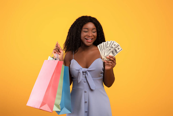 Winkelbudget. Portret van jonge zwarte vrouw houden fan van geld en kleurrijke shopper tassen over oranje achtergrond - Foto, afbeelding