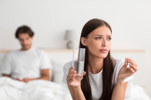 triste joven europeo dama celebrar píldora y vaso de agua, marido esperando esposa en cama en dormitorio - Foto, Imagen