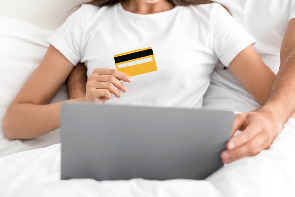 Kaukasischer Millennial-Mann umarmt Frau, Dame hält Kreditkarte und bestellt Waren auf Laptop, sitzt im Bett, beschnitten - Foto, Bild