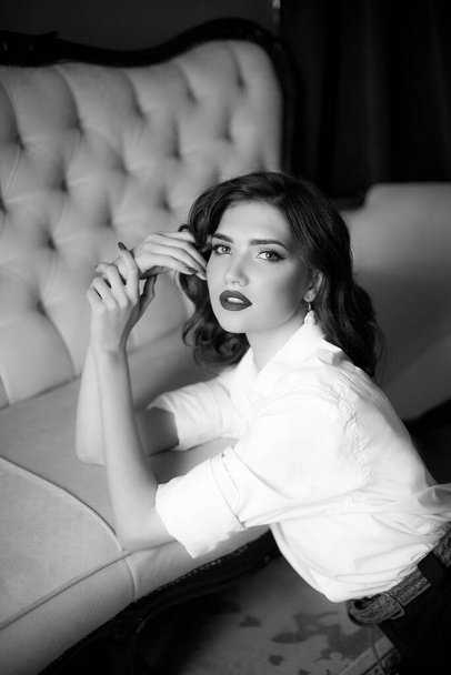 Attractive young girl with dark hair with Hollywood styling and makeup and red lipstick in a white men's shirt and dark trousers with a string of pearls around her neck in a classic interior. - Zdjęcie, obraz