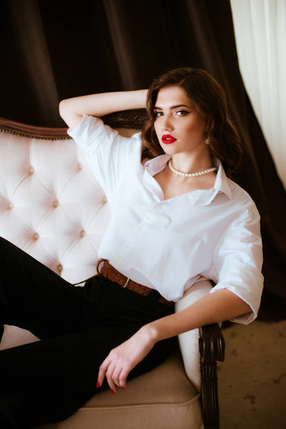 Attractive young girl with dark hair with Hollywood styling and makeup and red lipstick in a white men's shirt and dark trousers with a string of pearls around her neck in a classic interior. - Zdjęcie, obraz