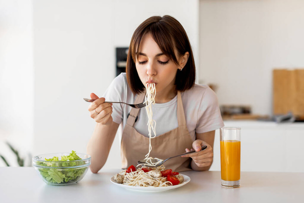 Młoda gospodyni domowa degustacja spaghetti i sałatki warzywnej, delektując się pysznym obiadem w domu, siedząc w nowoczesnej kuchni - Zdjęcie, obraz