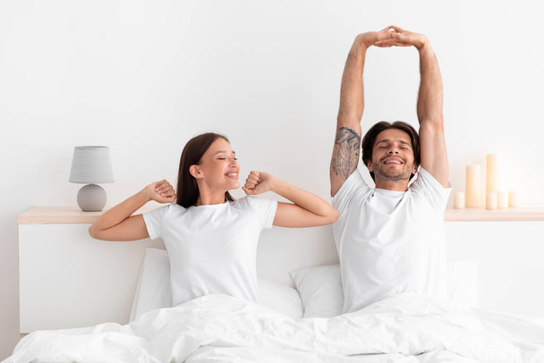 Happy caucasian millennial wife and husband woke up and stretching body sitting on bed in white bedroom - Fotoğraf, Görsel