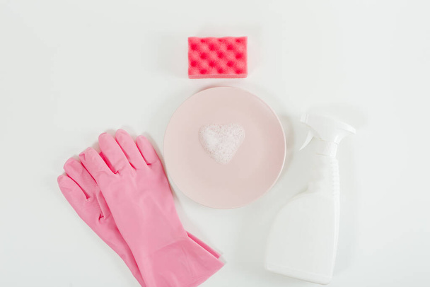 食器洗浄コンセプト。ピンクの手袋、プレート上のハート型の泡と白いスプレーボトル、赤いスポンジ。白地だ。平面、コピースペース - 写真・画像