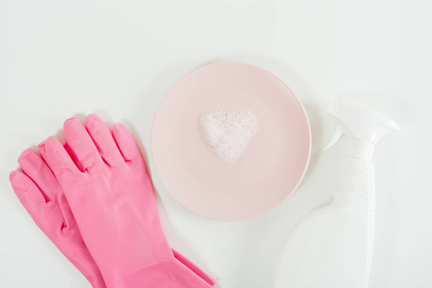 Mycie naczyń. Różowe rękawiczki, pianka w kształcie serca na talerzu i butelka z białym sprayem. Białe tło. Płaskie ułożenie, przestrzeń kopiowania - Zdjęcie, obraz
