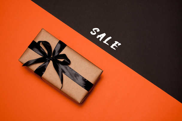 Een rechthoekige geschenkdoos verpakt in kraftpapier, versierd met een zwart satijnen lint en strik, ligt op een oranje-zwarte achtergrond. Verkoop. Zwarte vrijdag. Vlakke plaat. - Foto, afbeelding