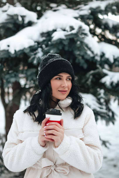 A beautiful young girl in warm winter clothes with a cup of hot coffee in her hands on a snowy winter walk in nature. Winter girl concept. - Zdjęcie, obraz