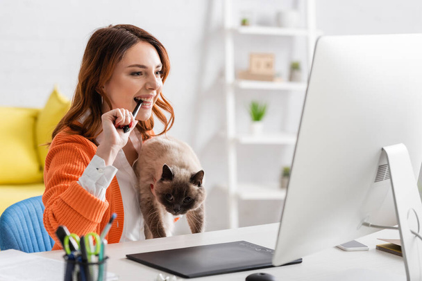 ενθουσιασμένος σχεδιαστής κρατώντας γάτα και δάγκωμα stylus, ενώ εργάζονται στο σπίτι - Φωτογραφία, εικόνα
