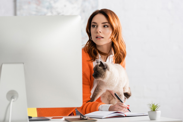 fiatal nő gazdaság macska és írás notebook miközben nézi elmosódott számítógép monitor - Fotó, kép