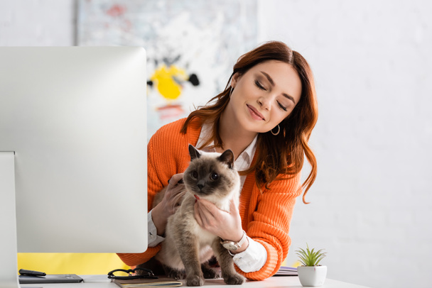 mujer sonriente abrazando gato en escritorio cerca de monitor de computadora - Foto, Imagen