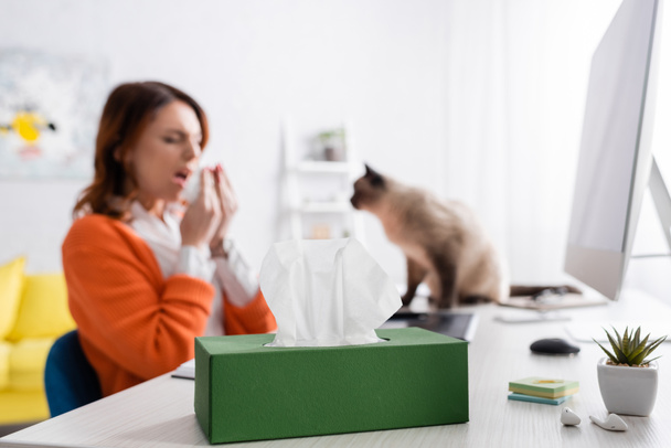 verschwommene allergische Frau niest in der Nähe von Katze und Packung Papierservietten auf Schreibtisch - Foto, Bild