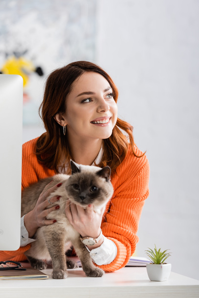 cheerful woman looking away while hugging cat sitting on desk - Фото, изображение