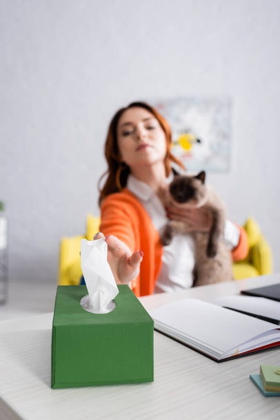 verschwommene allergische Frau nimmt Papierserviette, während sie mit Katze am Schreibtisch sitzt - Foto, Bild