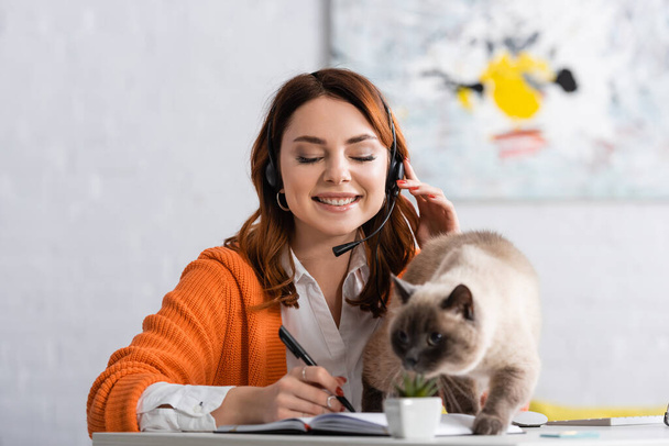mujer feliz en la escritura de auriculares en el cuaderno cerca del gato borroso en el escritorio - Foto, imagen