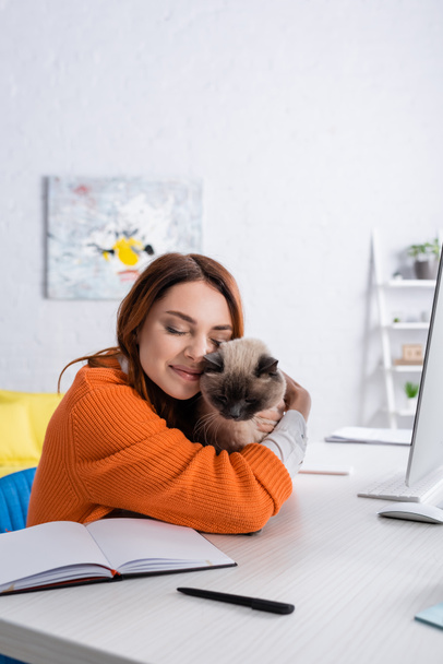 happy woman embracing cat while sitting near work desk at home - Фото, зображення