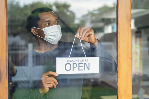 African american business man with face mask holding the sign for the reopening and looking away of the place after lockdown quarantine. Small business owner reopen and back to new normal concept. - Photo, image