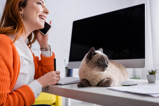 Счастливый фрилансер разговаривает по мобильному телефону у монитора с экраном и кошкой на рабочем столе - Фото, изображение