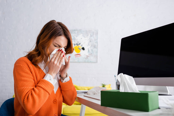 mujer enferma con los ojos cerrados estornudos en servilleta de papel cerca del monitor de la computadora con pantalla en blanco - Foto, Imagen