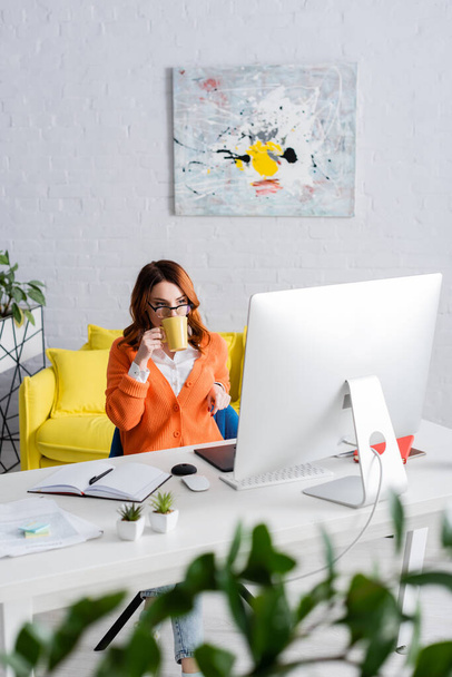 giovane designer in occhiali che beve tè mentre lavora vicino al monitor del computer a casa - Foto, immagini