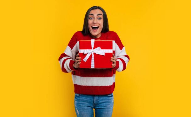 Close up photo of attractive smiling happy excited young brunette woman with gift box in hands isolated on yellow background - Fotó, kép