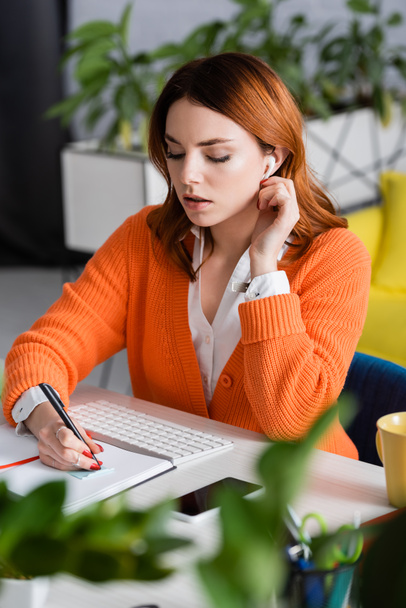 woman in earphone writing in notebook near keyboard and smartphone on blurred foreground - Foto, immagini