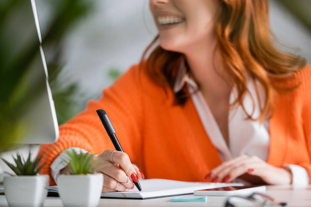 bijgesneden weergave van wazig vrouw schrijven in notebook in de buurt van computer monitor en smartphone op het bureau - Foto, afbeelding