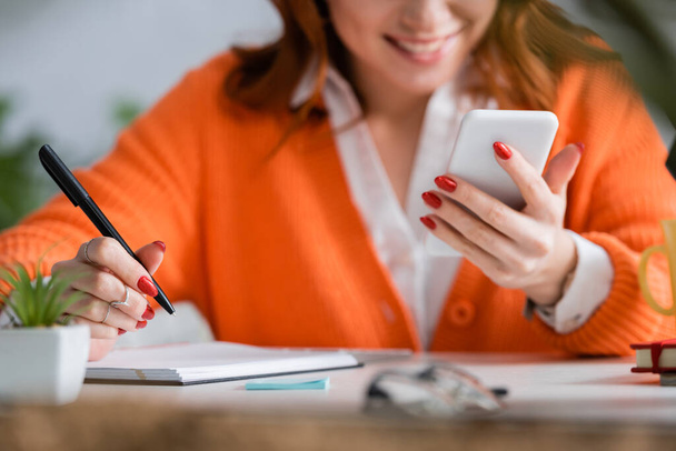 oříznutý pohled rozmazané ženy držící pero a mobilní telefon při práci doma - Fotografie, Obrázek