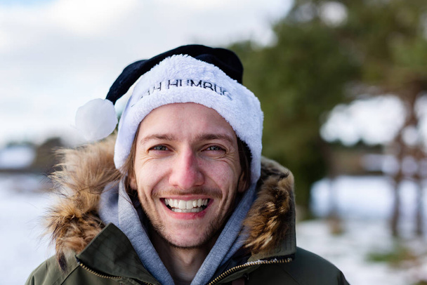 Ein ironisches Porträt eines lächelnden und glücklichen jungen Mannes, der einen schwarzen Weihnachtsmannhut mit der Aufschrift Bah Humbug trägt. - Foto, Bild