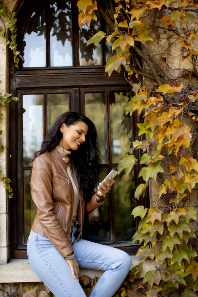 Όμορφη νεαρή γυναίκα που χρησιμοποιεί το κινητό τηλέφωνο, ενώ κάθεται στο παράθυρο σφραγίδα το φθινόπωρο - Φωτογραφία, εικόνα