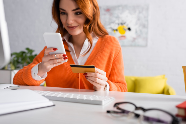 wazig vrouw glimlachen terwijl het houden van credit card in de buurt van toetsenbord thuis  - Foto, afbeelding