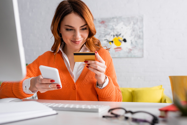 donna sorridente in possesso di carta di credito e smartphone mentre lavora a casa - Foto, immagini