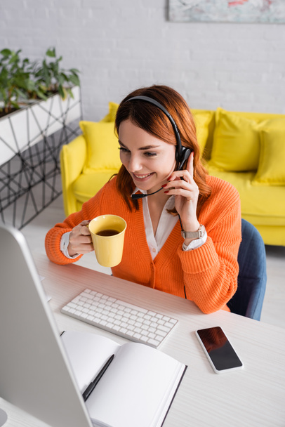 glimlachende vrouw in headset houden kopje thee tijdens het werken in de buurt wazig computer thuis - Foto, afbeelding