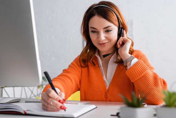 smiling woman in headset writing in notebook while working at home - Photo, Image