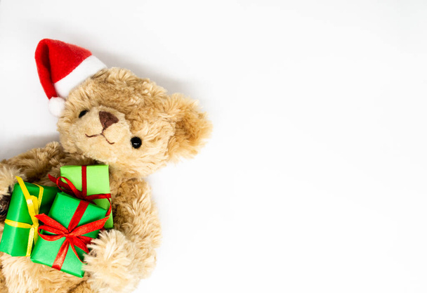 A stuffed toy Teddy bear in a red Santa Claus hat with a pompom on one ear, holding green gift boxes in its paws. White background, copy space. The concept of Christmas gifts, sales. - Фото, изображение