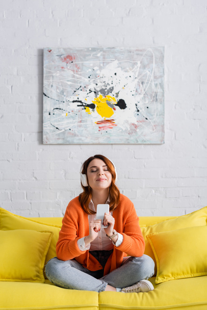 pleased woman with closed eyes listening music while sitting on yellow couch with crossed legs - Photo, image