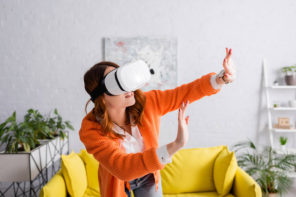 young woman in vr headset gesturing with outstretched hands while gaming in living room - Фото, изображение