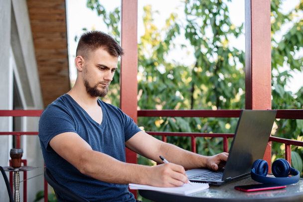 Un jeune homme barbu utilise un ordinateur portable pour travailler à l'extérieur, travailler projet indépendant en ligne à l'extérieur à la maison, beau étudiant étudiant, apprendre, éducation en ligne, pigiste - Photo, image