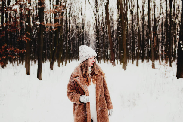 Zdjęcie młodej pięknej kobiety w futrze stojącej na białym śniegu w zimowym lesie. Dziewczyna jest uśmiechnięta i szczęśliwa z zamkniętymi oczami. Kobieta ma ferie zimowe, szczęśliwie czeka na święta Bożego Narodzenia - Zdjęcie, obraz