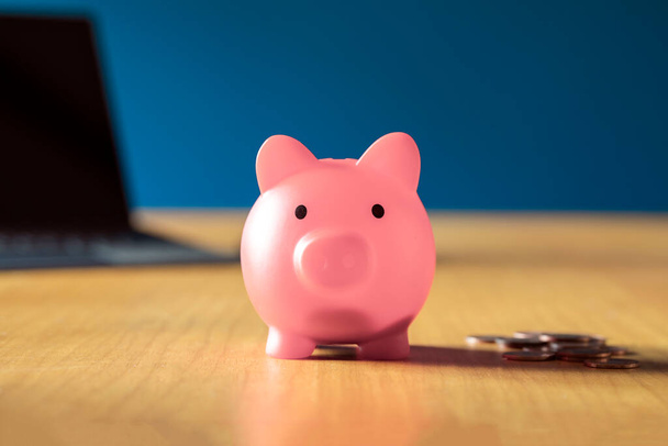 Розовый поросенок на деревянном офисном столе дома с грудой монет, сбережения, инвестиционный бизнес и финансовая концепция рядом - Фото, изображение