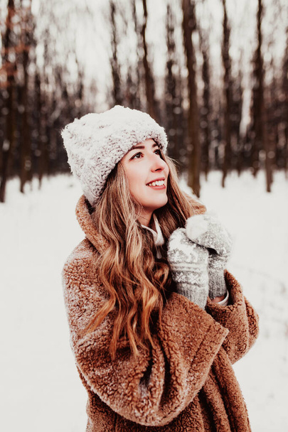 Hermosa mujer joven en el bosque de invierno de lujo nevado. Chica con guantes esponjosos, gorra y abrigo. Bosque de Navidad, árboles sobre fondo borroso. Cruzó las manos debido al clima frío. Foto de alta calidad - Foto, Imagen